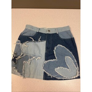 Peut inclure: Une mini-jupe en jean au design patchwork composé de différentes nuances de bleu. La jupe présente une application en forme de cœur effiloché sur le côté droit et des panneaux de tissu effilochés sur le côté gauche, avec une fermeture à bouton.