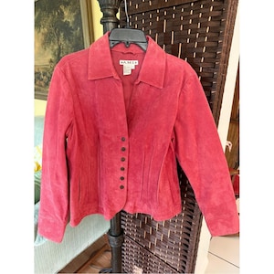 May include: A vibrant pink suede jacket featuring a classic collar, long sleeves, and a front button closure with six dark circular buttons. The garment includes vertical seam detailing along the front and a label at the neck reading A.M.I.