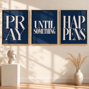 Può includere: Tre stampe rettangolari da parete con cornici dorate, caratterizzate da uno sfondo blu scuro con testo color crema. La prima stampa riporta PRAY, la seconda UNTIL SOMETHING e la terza HAPPENS. Le stampe sono disposte orizzontalmente su una parete.