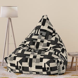 May include: A large beanbag chair with a geometric black, white, and gray pattern. The chair is triangular and sits on a patterned rug. A white lamp and small side table are in the background.