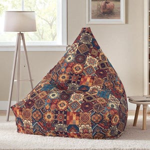 May include: A large, patterned beanbag chair with a patchwork design. The chair features a mix of geometric and floral patterns in shades of red, blue, orange, and brown. It is placed on a white rug in a living room setting.