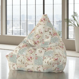 May include: A beanbag chair with a patchwork design featuring floral patterns in pink, cream, and pale blue. The chair is triangular and positioned on a light-coloured floor. A large window with a city view is in the background.