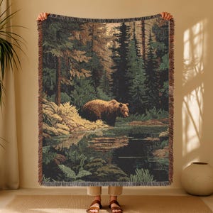 May include: A woven throw blanket featuring a forest scene with a brown bear standing near a lake. The blanket has a detailed design with shades of green, brown, and tan. The edges are finished with a fringe.