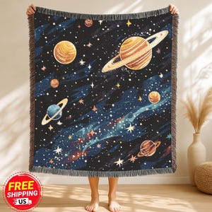 May include: A woven throw blanket featuring a celestial design with planets, stars, and a galaxy pattern in shades of blue, gold, and brown. The blanket has a fringed edge and is held up by a person.