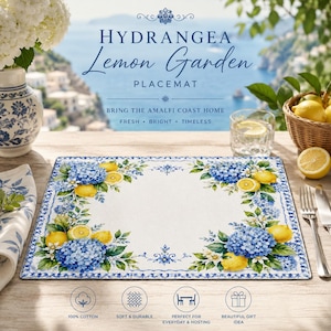 Puede incluir: Un mantel individual rectangular con fondo blanco, adornado con flores de hortensia azules y limones amarillos. El mantel individual tiene un borde azul y el texto "HYDRANGEA Lemon Garden PLACEMAT". También se ve una cesta de limones y un vaso de agua.