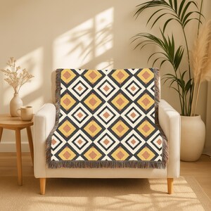 May include: A throw blanket with a geometric diamond pattern in black, white, yellow, and brown. The blanket is draped over a white armchair. The edges of the blanket have a fringe.