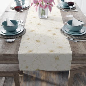 May include: A cream-coloured table runner with a floral design, set on a wooden table. The runner features a repeating pattern of large, stylised flowers in gold. The table is set with teal-coloured bowls, plates, and wine glasses.