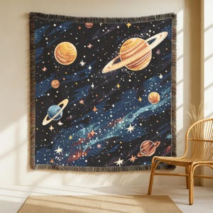 Op de afbeelding: Een geweven wandtapijt met een ruimte-thema ontwerp, met planeten, sterren en een melkwegstelsel op een donkerblauwe achtergrond. De planeten zijn oranje, bruin en blauw, met witte sterren. Het wandtapijt heeft een gefranjerde rand.