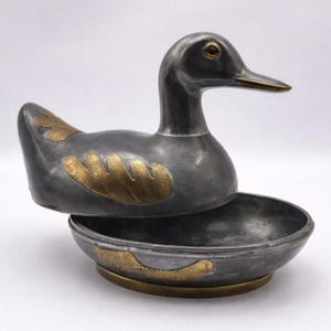 Könnte beinhalten: Eine zinnfarbene, entenförmige Schmuckschatulle mit Deckel. Die Ente hat goldfarbene Akzente auf ihren Flügeln und um den Boden der Schale. Auch der Schnabel und die Augen sind goldfarben. Der Deckel ist geöffnet und gibt die darunter liegende Schale frei.