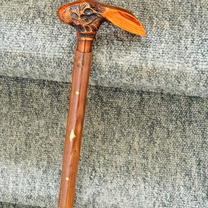 Puede incluir: Un bastón de madera con un mango tallado en forma de cabeza de conejo. El mango presenta tonos marrones y anaranjados, con tallado detallado. El eje es marrón pulido con elementos decorativos incrustados. Un accesorio único y funcional.