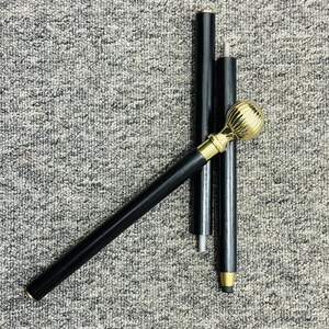 Op de afbeelding: Een zwarte wandelstok met een goudkleurige, bolvormige handgreep en gouden accenten. De wandelstok is gedemonteerd, met de handgreep en drie zwarte secties zichtbaar. De achtergrond is een grijs, getextureerd oppervlak.