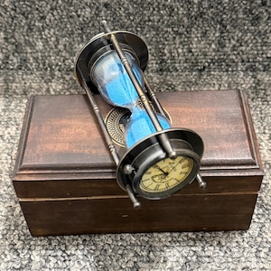 May include: An antique-style hourglass with blue sand, resting on a wooden box. The hourglass features a metal frame and a small clock face. The box is dark brown, and the background is a textured grey.