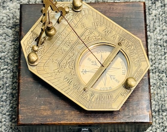 Puede incluir: Un reloj de sol de latón antiguo con brújula, en una caja de madera. El reloj de sol presenta números romanos, un gnomon y una rosa de los vientos que indica las direcciones. La caja es de color marrón oscuro.