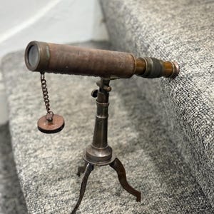 Könnte beinhalten: Ein Vintage-Teleskop aus Messing und Holz auf einem Stativ. Das Teleskop hat einen braunen Holzschaft und eine kleine Linse. Eine kleine, runde Messingkappe hängt an einer Kette, die am Teleskop befestigt ist. Das Stativ hat drei gebogene Beine.