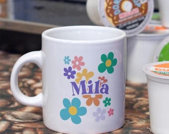 May include: A white ceramic mug featuring a colorful floral pattern and the name Mila printed in purple letters. Several coffee pods are arranged in the background, including brands like The Original Donut Shop, Green Mountain, and Dunkin.