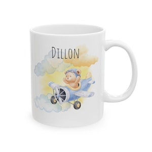 May include: White ceramic mug featuring a watercolor illustration of a teddy bear in a blue airplane, with the name "Dillon" above. The design includes yellow and blue clouds. The mug has a white handle.