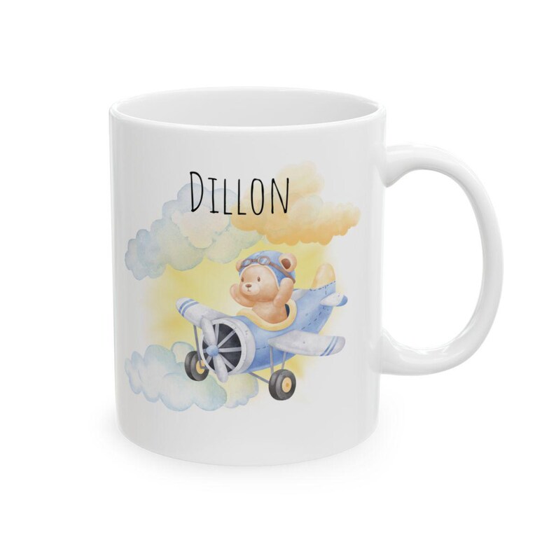 May include: White ceramic mug featuring a watercolor illustration of a teddy bear in a blue airplane, with the name "Dillon" above. The design includes yellow and blue clouds. The mug has a white handle.