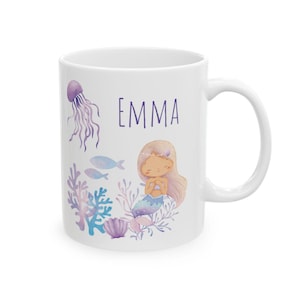 May include: White ceramic mug featuring a watercolor illustration of a mermaid, jellyfish, fish, and coral. The name "EMMA" is printed in purple. The mug has a curved handle.