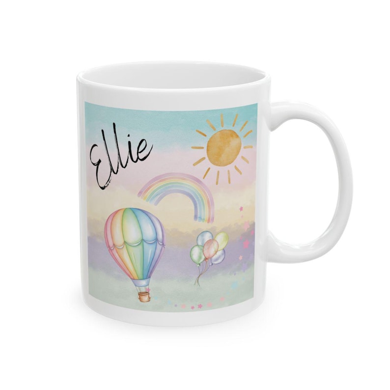 May include: White ceramic mug featuring a colorful design with a hot air balloon, rainbow, sun, and balloons. The name "Ellie" is written in cursive. The mug has a curved handle and is suitable for hot or cold beverages.