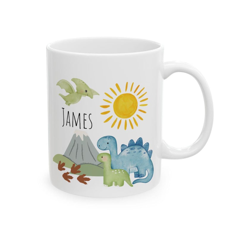 May include: White ceramic mug featuring a watercolor design with a green Pterodactyl, a yellow sun, and two dinosaurs. The name "JAMES" is printed in black. The mug has a curved handle.