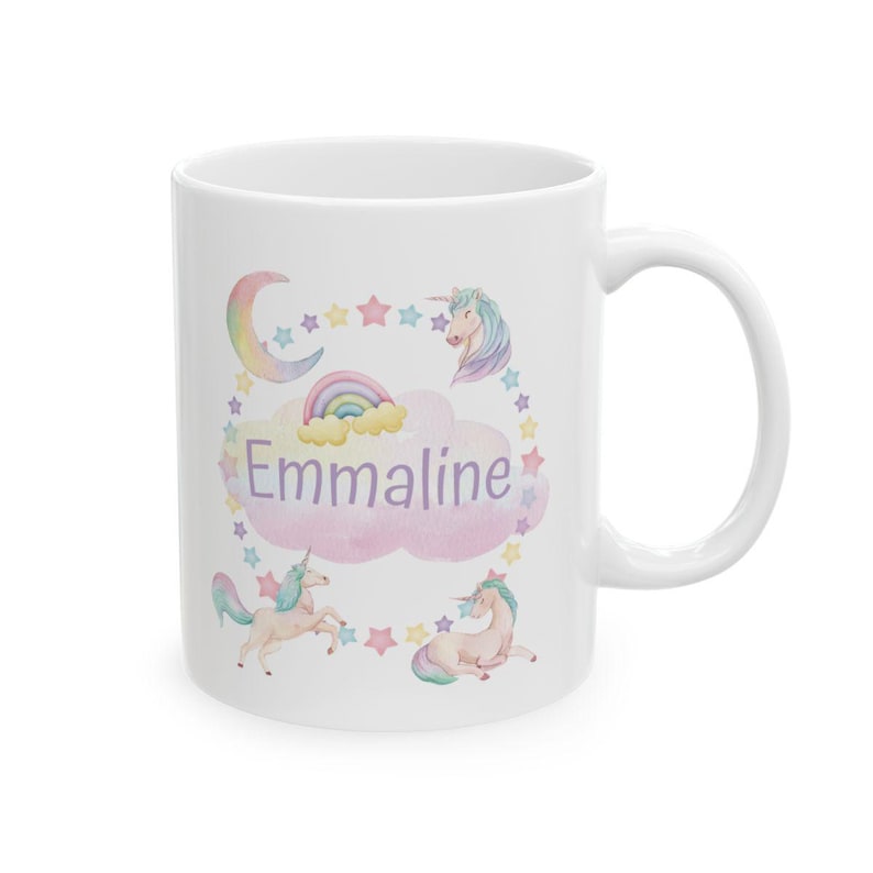 May include: White ceramic mug featuring a whimsical design with unicorns, a rainbow, and stars. The name "Emmaline" is displayed in a pink cloud. The mug has a curved handle.