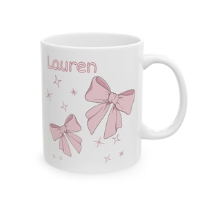 May include: White ceramic mug featuring the name "Lauren" in pink script, along with two pink bows and star accents. The mug has a curved handle and a glossy finish.