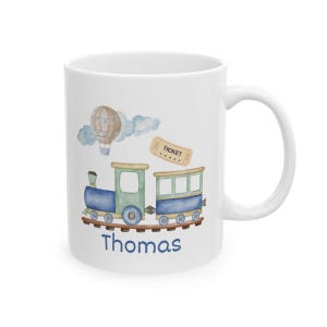 May include: White ceramic mug featuring a watercolor illustration of a blue and green train, a hot air balloon, clouds, and a ticket. The name "Thomas" is written in blue script below the train. The mug has a white handle.