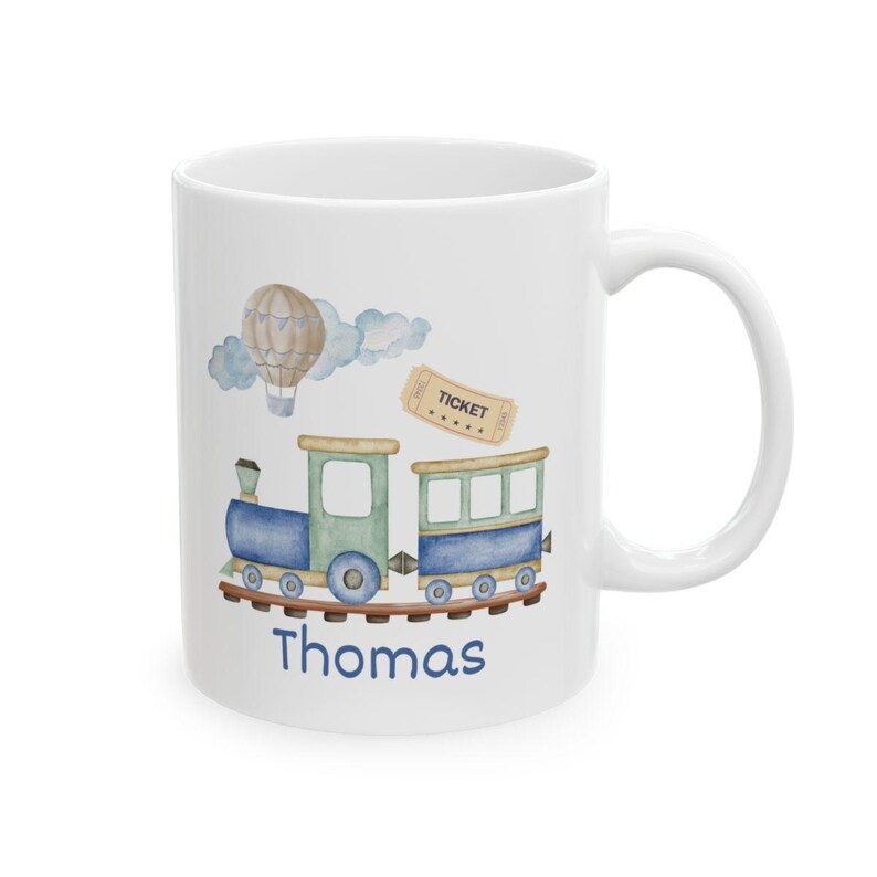 May include: White ceramic mug featuring a watercolor illustration of a blue and green train, a hot air balloon, clouds, and a ticket. The name "Thomas" is written in blue script below the train. The mug has a white handle.