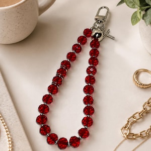 May include: A wristlet keychain featuring a loop of faceted, translucent red beads separated by small silver-toned spacers. The keychain includes a silver-toned lobster clasp, a smooth silver-toned spherical bead, and a small silver-toned cross charm.