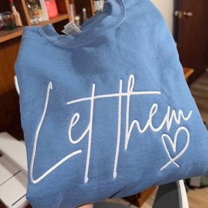 以下が含まれることがあります： 白い筆記体で「Let them」と小さなハートが描かれた、水色のスウェットシャツ。スウェットシャツは折りたたまれており、テキストは前面の中央に配置されています。