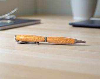 May include: A ballpoint pen with a polished light brown wood barrel featuring a visible grain pattern. The pen includes a dark metallic tip, a matching center band, and a sturdy pocket clip attached to the cap, all finished in a dark gunmetal tone.