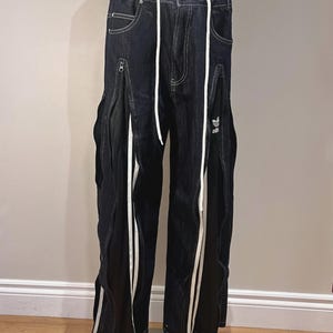 May include: Dark wash denim trousers with a white drawstring and white side stripes. Features side zips and the Adidas logo on the right leg. The trousers have a relaxed fit and a unique design.