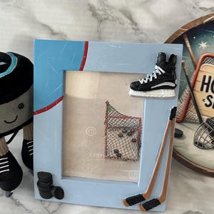 May include: A light blue picture frame with hockey-themed decorations. The frame includes a hockey skate, hockey sticks, and a stack of pucks. The image inside the frame shows a hockey net and pucks.