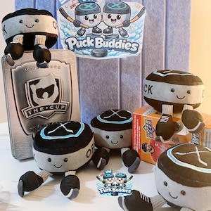 Peut inclure: Plusieurs personnages de palets de hockey en peluche avec des visages souriants, des gants et des chaussettes. Les personnages sont noirs, gris et blancs, avec un accent bleu. L'image comprend également une boîte de cartes de hockey et un logo "Puck Buddies".
