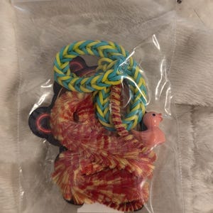 May include: A clear plastic bag containing a red and yellow fuzzy bear-shaped item, a blue and yellow rubber band bracelet, and a small pink plastic duck. The items are on a white, fuzzy surface.