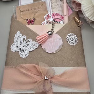 May include: A gift set with a brown paper envelope tied with a pink ribbon and a decorative bow. The set includes a butterfly necklace, a keychain with a pink tassel, and a white lace butterfly.