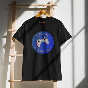 May include: Black t-shirt with a blue and purple circle graphic featuring a video game controller. The text "LIFE IS JUST ANOTHER LEVEL" is printed around the controller. The shirt is hanging on a wooden hanger.