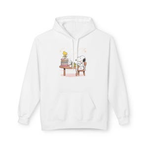 May include: White hoodie featuring a graphic of Snoopy sitting at a table with books and Woodstock. The illustration is in the center of the hoodie. The hoodie has a drawstring hood and a front pocket.