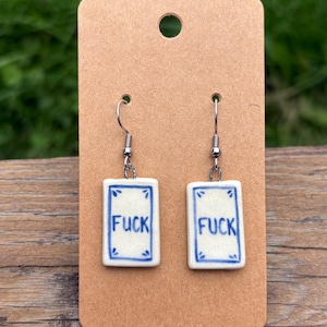 May include: A pair of ceramic earrings with the word "FUCK" in blue lettering on a white background. The earrings are rectangular with a blue border and hang from silver-colored hooks. They are displayed on a brown card.