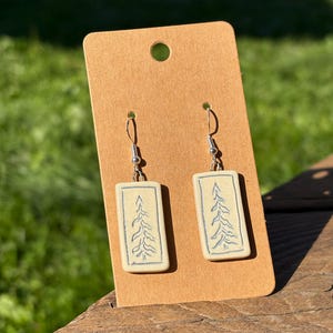 May include: A pair of rectangular ceramic earrings with a hand-drawn tree design in blue on a cream background. The earrings are displayed on a brown card with a hole at the top, showcasing a nature-inspired theme.