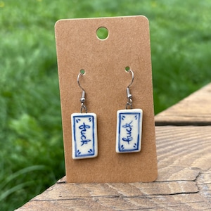 May include: A pair of rectangular ceramic earrings with the word "fuck" written in blue script. The earrings are white with a blue border and are displayed on a brown earring card.