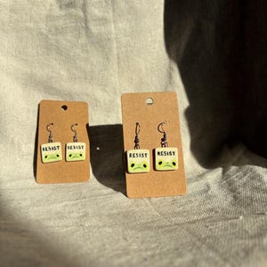 May include: Two pairs of earrings on brown cardboard earring cards. Each earring is a small, square, off-white tile with the word "RESIST" in black and a green face with black eyes. The earrings have silver-colored hooks.