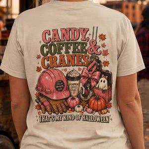 May include: A cream-colored short-sleeve t-shirt featuring a graphic on the back with the text CANDY, COFFEE & CRANES and THAT'S MY KIND OF HALLOWEEN. The design includes a pink hard hat, a coffee cup, a crane hook, a ghost, and pumpkins.