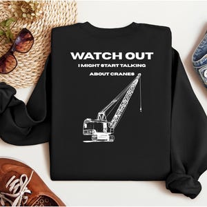 Może przedstawiać: Czarna bluza z białym napisem "WATCH OUT I MIGHT START TALKING ABOUT CRANES" i białą ilustracją żurawia. Bluza ma okrągły dekolt.