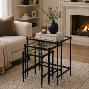 May include: A set of three nesting tables with black metal frames and clear glass tops. The tables are stacked together, featuring slender legs with a ribbed texture. The top table holds a black ribbed vase with greenery, a candle, and two books.
