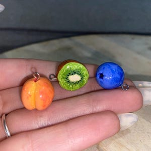 Pode incluir: Três pingentes de frutas em miniatura: um pêssego laranja, uma fatia de kiwi e um mirtilo azul. Cada pingente tem um laço prateado para prender a joias. O kiwi é verde com sementes pretas e um centro branco.