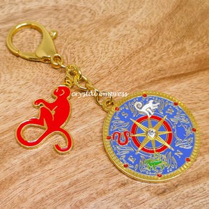 Puede incluir: Un llavero dorado con un dije de mono rojo y un dije de brújula azul y dorado. El dije de la brújula presenta un mono blanco, una serpiente roja y un diseño de estrella dorada. El llavero tiene un cierre de mosquetón.