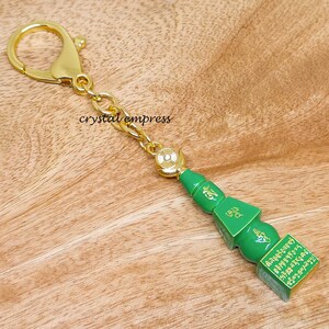 Peut inclure: Un porte-clés vert et or représentant une pagode miniature avec des inscriptions dorées. Le porte-clés est doté d'une chaîne dorée et d'un anneau brisé doré.