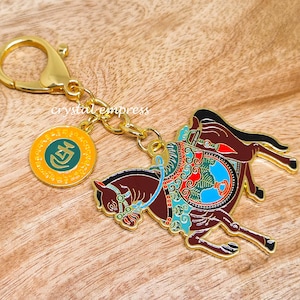 Puede incluir: Llavero dorado con un caballo esmaltado detallado con una silla de montar colorida y un dije redondo con la palabra "Celi" en letras verdes.