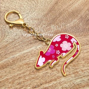 May include: A gold-coloured keyring featuring a red rat design with pink floral accents. The rat is outlined in gold and attached to a gold clasp and chain. The text "crystal empress" is visible.